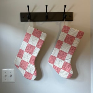 May include: Two red and white checkered Christmas stockings hanging on a brown wooden hook rack.