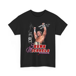 Könnte beinhalten: Schwarzes T-Shirt mit einer Grafik von Shawn Michaels, einem professionellen Wrestler, mit erhobenen Armen. Das Design enthält den Text "Shawn Michaels" in rosa Schreibschrift und "The Heartbreak Kid" in Rot.