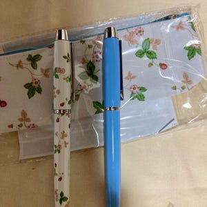 以下が含まれることがあります： 2本のペン。1本は花柄の白、もう1本は水色で、お揃いのペンケースの隣にあります。白いペンにはイチゴと花の模様があります。ペンケースにも同様の花柄があります。