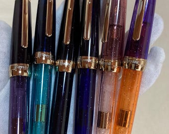 Peut inclure: Six stylos-plume avec des corps translucides à paillettes en violet, bleu sarcelle, noir, bleu, rose et orange. Chaque stylo présente un clip doré, une bague dorée et le mot "Fonte" gravé sur la bague du capuchon avec une finition métallique.