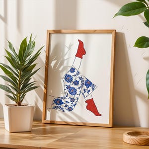 Puede incluir: Una impresión artística enmarcada que presenta un dibujo lineal de piernas y pies. La persona lleva calcetines rojos y pantalones con estampado azul y blanco. La obra de arte está en un marco de madera y descansa sobre una superficie de madera.