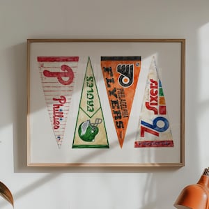 Könnte beinhalten: Ein gerahmter Wanddruck mit vier dreieckigen Sportwimpeln. Die Wimpel zeigen die Logos und Namen der Philadelphia Phillies, Eagles, Flyers und 76ers in den Farben Rot, Grün, Orange und Blau auf einem weißen Hintergrund.
