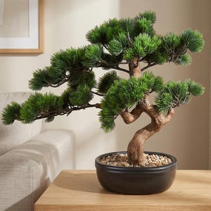 May include: A small, artificial bonsai tree in a black ceramic pot. The tree has vibrant green foliage and a brown, curved trunk. The pot is filled with small, light-colored pebbles. The bonsai is displayed on a light-colored wooden surface.