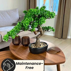 Puede incluir: Un peque&ntilde;o bons&aacute;i artificial con follaje verde vibrante y un tronco marr&oacute;n texturizado en una maceta negra ovalada. Dos candelabros marrones y una pila de posavasos marrones est&aacute;n sobre una mesa de madera. La mesa tiene el texto "Maintenance-Free".
