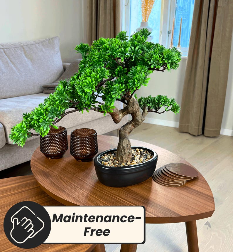 Puede incluir: Un peque&ntilde;o bons&aacute;i artificial con follaje verde vibrante y un tronco marr&oacute;n texturizado en una maceta negra ovalada. Dos candelabros marrones y una pila de posavasos marrones est&aacute;n sobre una mesa de madera. La mesa tiene el texto "Maintenance-Free".