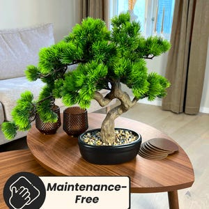 May include: A small, artificial bonsai tree with vibrant green foliage and a textured, brown trunk sits in a black pot filled with small stones. The tree is on a wooden table with the text "Maintenance-Free".