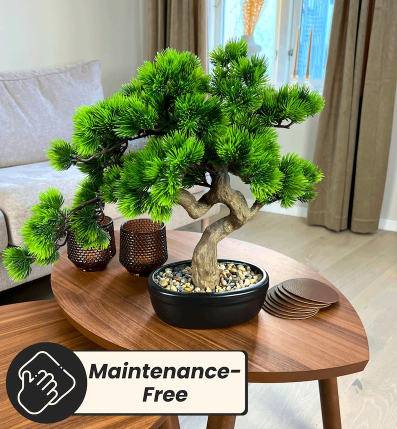 May include: A small, artificial bonsai tree with vibrant green foliage and a textured, brown trunk sits in a black pot filled with small stones. The tree is on a wooden table with the text "Maintenance-Free".