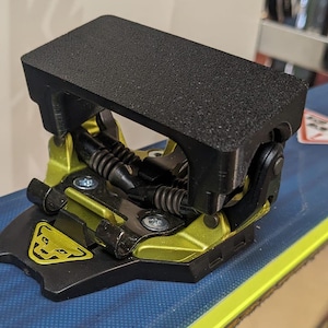 May include: A black ski binding with a black and yellow release mechanism. The binding is mounted on a blue and yellow ski.