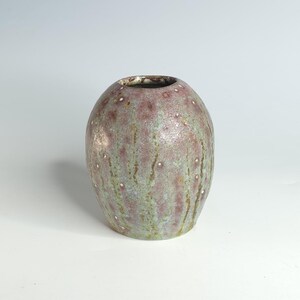 May include: A small, round ceramic vase with a speckled, textured surface. The vase features a blend of muted green and mauve colors with vertical streaks and small white dots. The opening is dark, contrasting with the lighter body.