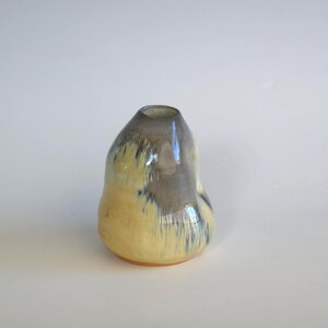 May include: A handcrafted ceramic vase with a unique, organic shape. The vase features a gradient of colors, transitioning from a pale yellow base to a gray top with blue accents. The glossy finish enhances the visual appeal.