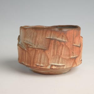 May include: A handmade, textured ceramic bowl with a reddish-brown hue. The bowl features a unique, carved design with a rough, organic texture. The base is a lighter shade of brown, and the rim has a slightly uneven edge.