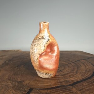 May include: A handcrafted ceramic vase with a unique, organic shape. The vase features a blend of warm colors, including shades of orange, beige, and brown. The textured surface adds visual interest, making it a decorative piece for any home. The vase is approximately 8 inches tall.