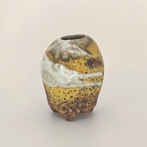 May include: A small, handcrafted ceramic vase with a unique, organic shape. The vase features a speckled, yellow base that transitions to a white and brown upper section. It has a small opening and three small feet.