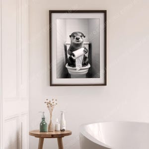 May include: A black and white photo of an otter sitting on a toilet holding a roll of toilet paper. The otter is looking directly at the camera.