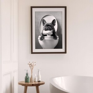 May include: Black and white photo of a French bulldog sitting in a toilet bowl, holding a roll of toilet paper in its mouth. The photo is framed in a dark brown frame.