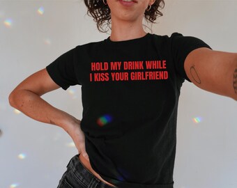 Op de afbeelding: Zwart T-shirt met rode tekst die "Hold my drink while I kiss your girlfriend." zegt.
