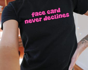 Op de afbeelding: Zwart T-shirt met de tekst "face card never declines" in roze letters.