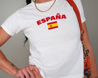 May include: White t-shirt with the word "ESPAÑA" in red letters and a small image of the Spanish flag.