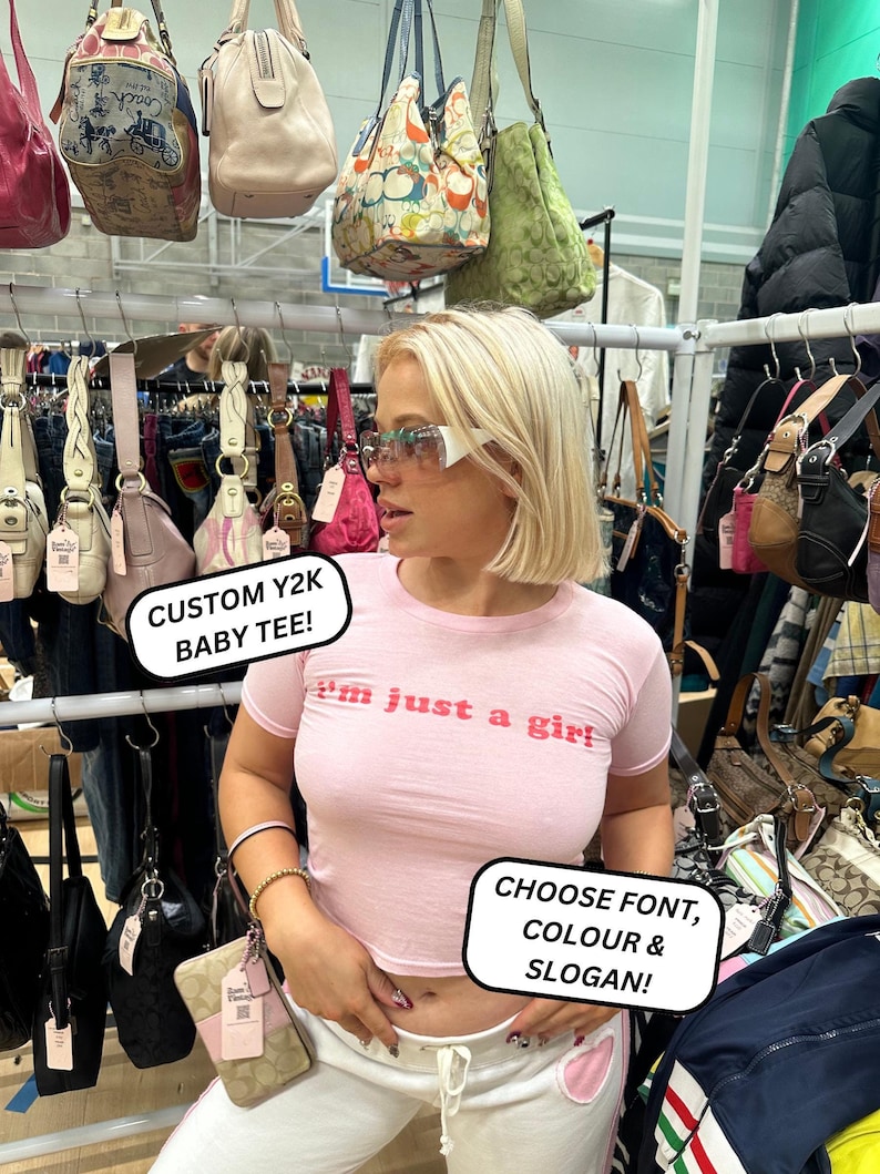 Peut inclure: Une femme portant un haut court rose avec le texte "I'm just a girl" se tient devant un pr&eacute;sentoir de sacs &agrave; main. Elle tient un petit sac &agrave; main brun avec une bandouli&egrave;re blanche. Le texte "CUSTOM Y2K BABY TEE!" et "CHOOSE FONT, COLOUR & SLOGAN!" est &eacute;crit sur des pancartes dans l'image.
