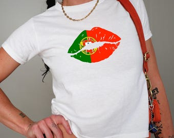 May include: White t-shirt with a graphic of lips in the colours of the Portuguese flag: red, green, and yellow. The top has a crew neck and short sleeves. A gold chain and a red shoulder bag are also visible.