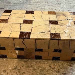 May include: A rectangular box with a checkered pattern made of light brown and dark brown pieces. The box is made of a material that looks like wood or stone.