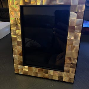 May include: A gold picture frame with a mosaic of iridescent shell tiles. The frame has a black mat and a clear glass front.