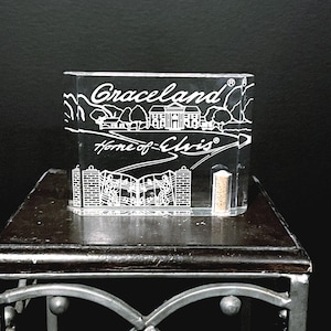 May include: A clear, rectangular Graceland souvenir. The front features the words "Graceland Home of Elvis" in white script and a detailed illustration of the mansion. The item is displayed on a dark wooden surface.