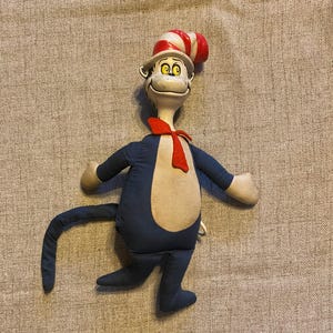 May include: A plush toy of the Cat in the Hat character. The figure is primarily navy blue with a white belly and arms. It wears a red bow tie and a red and white striped hat. The cat has a smiling face with yellow eyes.