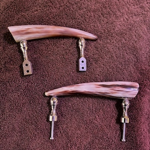 May include: Two cabinet handles with a curved, horn-like design in shades of brown and cream. The handles are attached to silver-colored metal bases with screw-in posts for installation. The handles are likely for furniture.