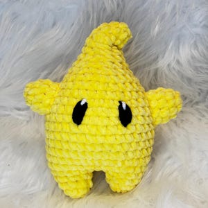 Puede incluir: Un juguete de peluche con forma de estrella de color amarillo brillante hecho de hilo grueso y suave. La estrella tiene una punta superior, dos brazos laterales pequeños y dos piernas cortas en la base. Tiene dos ojos negros ovalados con reflejos blancos.