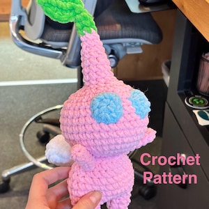 May include: A handmade crochet figure in pink, blue, green, and white. The figure has a bulbous body, blue eyes, and a green leaf-like structure on top. The text "Crochet Pattern" is visible.