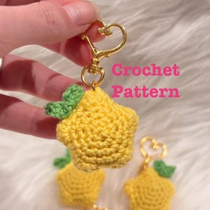 May include: A yellow star-shaped crochet keychain with green leaves attached to a gold-colored heart-shaped clasp. The text "Crochet Pattern" is printed in pink on the right side. The item is a small, handmade textile accessory for keys.
