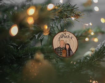 May include: A round wooden slice ornament featuring a hand-drawn illustration of two people and a small dog. The text "The Farahs" is written in black script at the top. The ornament has a natural wood grain texture and is hung by a thin string.