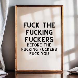 May include: A framed art print with the text "FUCK THE FUCKING FUCKERS BEFORE THE FUCKING FUCKERS FUCK YOU" in bold black letters. The frame is a warm brown color, and the background is a light beige.