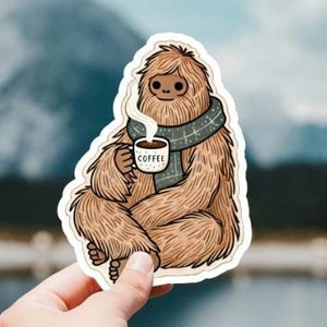 Może przedstawiać: Brązowe, futrzane stworzenie w zielonym szaliku trzymające biały kubek z napisem "COFFEE". Stworzenie siedzi ze skrzyżowanymi nogami i ma rozluźniony wyraz twarzy.