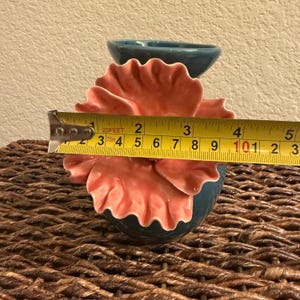 May include: A teal ceramic flower-shaped vase with coral-colored petals. The vase is approximately 4 inches wide. The vase is sitting on a woven surface. The vase has a small teal container on top.