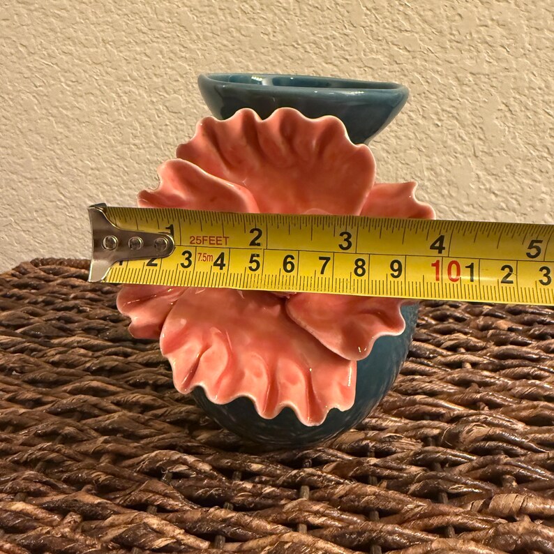 May include: A teal ceramic flower-shaped vase with coral-colored petals. The vase is approximately 4 inches wide. The vase is sitting on a woven surface. The vase has a small teal container on top.