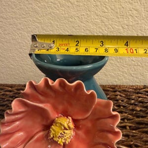 May include: A pink ceramic flower with a yellow center sits in front of a teal ceramic bowl. A yellow measuring tape is placed across the bowl, showing a measurement of approximately 3 inches.