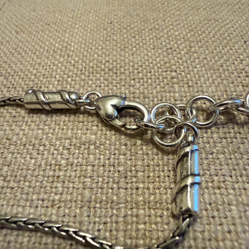 May include: A silver-toned metal necklace featuring a lobster claw clasp, a series of circular jump rings for length adjustment, and two cylindrical end caps with a diagonal grooved pattern attached to a woven chain link design.