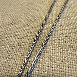 May include: A silver-toned wheat chain necklace featuring a tightly woven, braided metal link design. The chain has a polished finish with dark oxidized accents within the crevices of the links, creating a textured and detailed appearance.