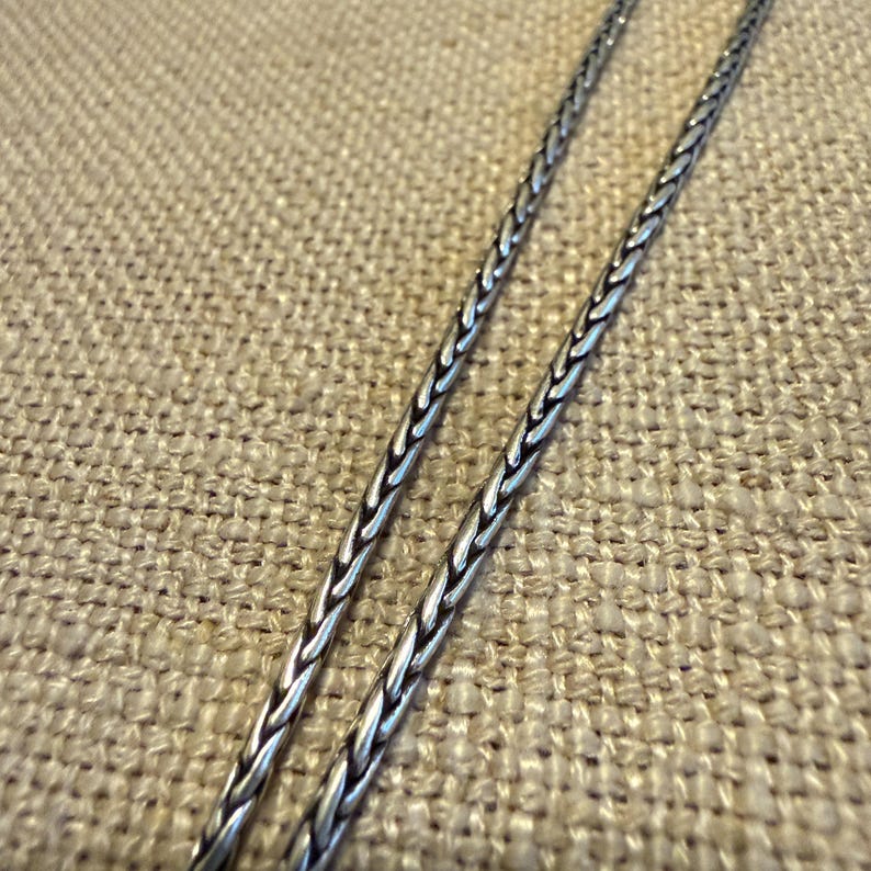 May include: A silver-toned wheat chain necklace featuring a tightly woven, braided metal link design. The chain has a polished finish with dark oxidized accents within the crevices of the links, creating a textured and detailed appearance.