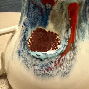 May include: Close-up of a handmade ceramic mug with a white base and a colorful glaze. The glaze features blue, red, and brown hues, creating a unique, artistic design. The mug has a handle and a textured surface.