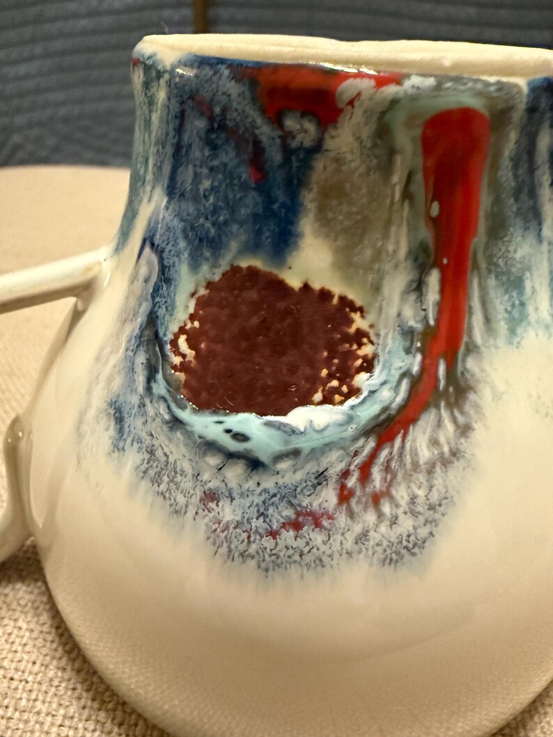 May include: Close-up of a handmade ceramic mug with a white base and a colorful glaze. The glaze features blue, red, and brown hues, creating a unique, artistic design. The mug has a handle and a textured surface.