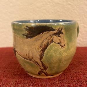 May include: A ceramic mug with a green and blue glaze features a hand-painted image of a white horse running. The mug has a crackled glaze and a rounded shape.
