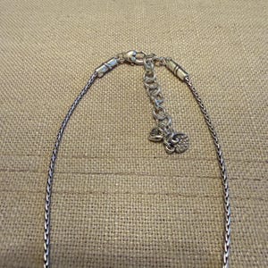 May include: A silver-toned chain necklace featuring a woven wheat-style link design. The necklace includes a lobster claw clasp, an adjustable extender chain with circular links, and two small heart-shaped charms attached to the end of the extender.