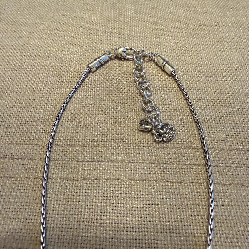 May include: A silver-toned chain necklace featuring a woven wheat-style link design. The necklace includes a lobster claw clasp, an adjustable extender chain with circular links, and two small heart-shaped charms attached to the end of the extender.