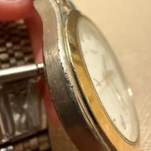 以下が含まれることがあります： シルバーカラーの金属製ケースとゴールドカラーのベゼルを備えたツートンカラーの腕時計。白い文字盤と金属製のリンクブレスレットが付いています。シルバーのケースの側面には、摩耗や表面の変色の跡がはっきりと見られます。