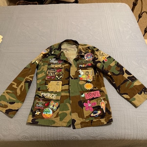 May include: Camouflage jacket with multiple patches. Patches include the words "FIERCE" and "LOVE", along with various designs such as a peace sign, a guitar, a sun, and a lightning bolt. The jacket is laid flat on a light blue bedspread.