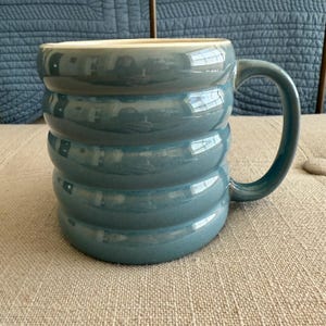 May include: A teal ceramic mug with a unique, ridged design. The mug has a large handle and a cream-colored interior. The exterior has a glossy finish. This mug is suitable for coffee, tea, or other beverages.