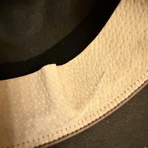 May include: Close-up of a hat's interior, showcasing the textured, light-colored lining and the dark, possibly felt, exterior. The stitching along the brim is visible, adding detail to the hat's construction. The hat appears to be a classic style.